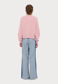 Άτομο με κοντά καστανά μαλλιά, που φοράει ροζ πουλόβερ και ευρύχωρο δίχρωμο τζιν παντελόνι με φαρδιές παντελόνες, στέκεται με την πλάτη προς το μέρος μας σε λευκό φόντο.