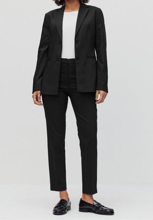 Femme portant un blazer noir sur mesure et un pantalon assorti, un haut blanc et des mocassins noirs brillants, debout devant un fond clair uni.