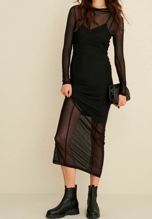Femme portant une robe noire longue transparente à manches longues avec une superposition en maille mi-longue, tenant une pochette noire et portant des bottines noires.