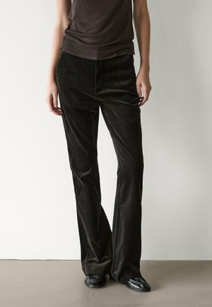 Personne portant un pantalon large marron foncé et des chaussures noires, debout devant un mur et un sol clairs et unis.