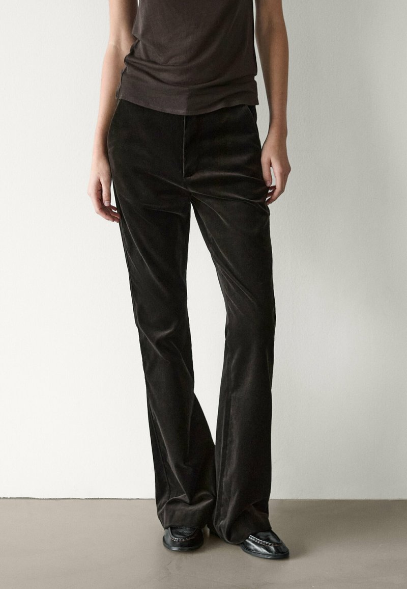 Personne portant un pantalon large marron foncé et des chaussures noires, debout devant un mur et un sol clairs et unis.