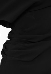 Abito nero con drappeggi laterali e una texture liscia. Il tessuto è leggero con pieghe morbide che creano una silhouette strutturata.
