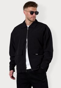 Black bomber jacket made of lightweight fabric, featuring a zip front, two front pockets, and a classic collar. Worn over a white shirt.