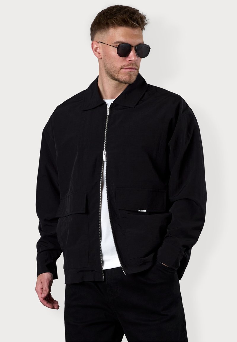 Black bomber jacket made of lightweight fabric, featuring a zip front, two front pockets, and a classic collar. Worn over a white shirt.