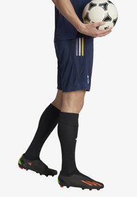 Jugador de fútbol con camiseta azul marino con rayas amarillas y moradas, pantalones cortos negros, calcetas negras y botas negras con acentos verdes y rojos, sosteniendo un balón de fútbol.