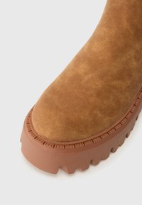 Bota de ante marrón con suela de goma gruesa y texturizada y puntera redondeada, fotografiada sobre un fondo blanco.
