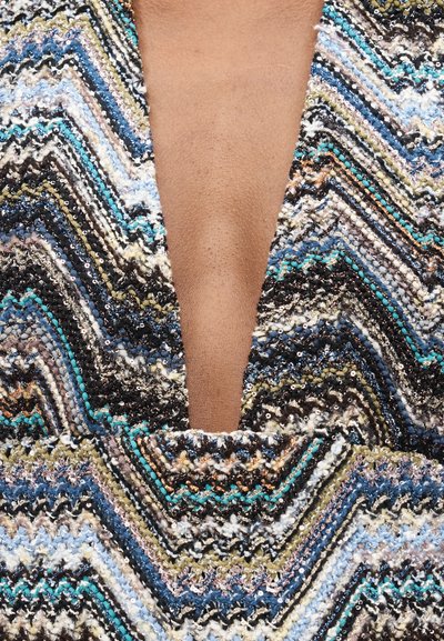 Κοντινό πλάνο σε ένδυμα με βαθύ V-λαιμόκοψη και πολύχρωμο zigzag πλεκτό μοτίβο σε μπλε, μαύρο, λευκό, μπεζ και τυρκουάζ με υφή νήματος και παγέτες.