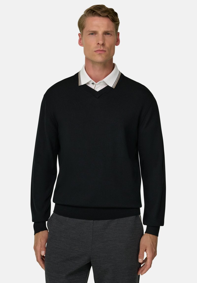 Homme portant un pull noir col en V par-dessus une chemise blanche à col avec un pantalon gris, debout devant un fond clair uni.