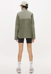 Pullover in poliestere felpato verde oliva con colletto alto, pannelli lisci a contrasto, maniche lunghe e vestibilità rilassata, abbinato a pantaloni corti neri e sneaker.