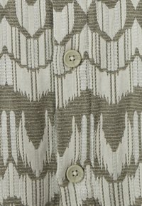 Geweven textiel met groene en witte geometrische vormen. Bevat een ronde, beige knop in het midden. Textuur van linnen.