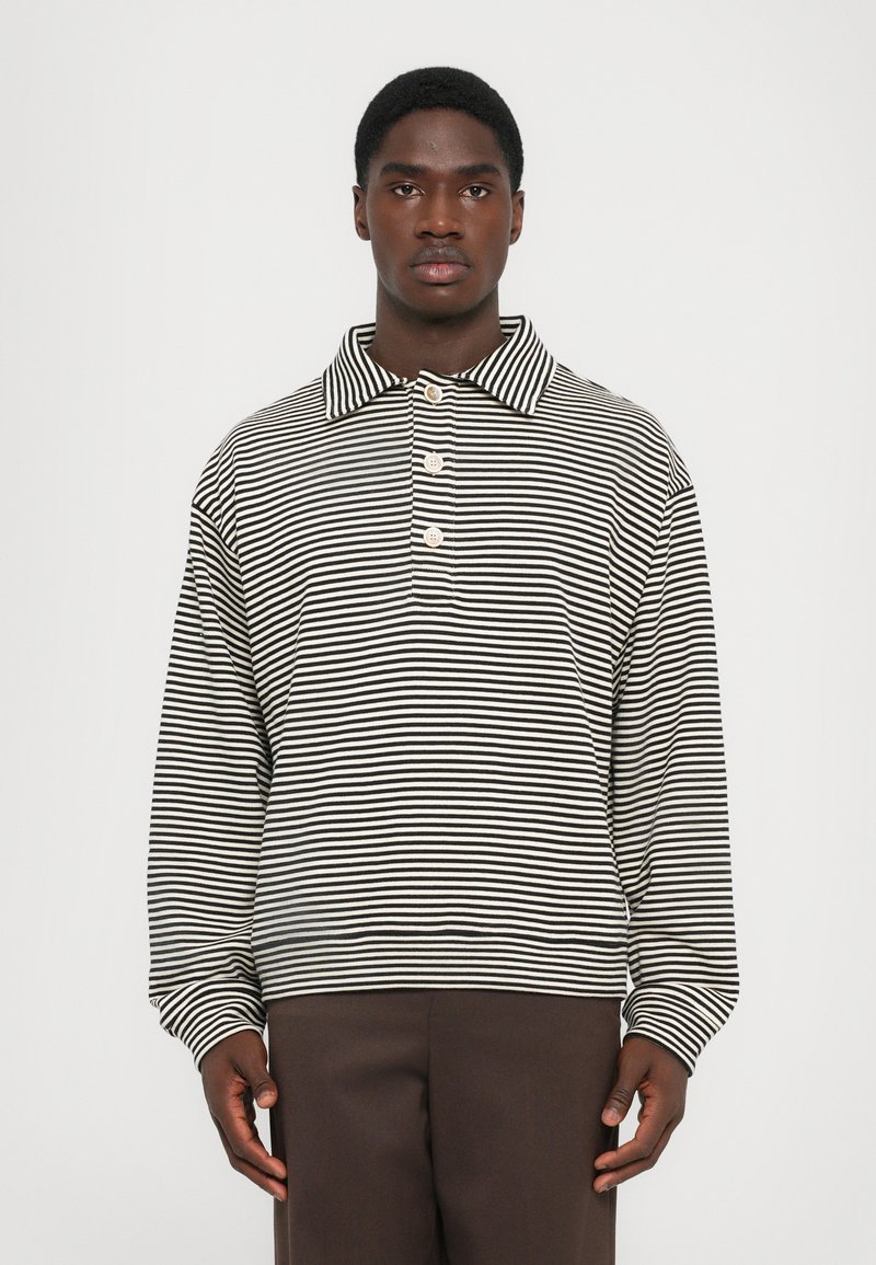 Homme portant un polo à rayures horizontales noir et blanc à manches longues avec trois boutons et un pantalon marron foncé uni.