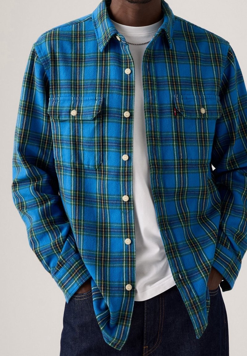 Homme portant une chemise à carreaux bleue et verte boutonnée sur un t-shirt blanc, les mains dans les poches d'un jean foncé.