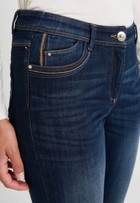 Jeans de denim azul oscuro con corte ajustado, con costuras doradas, cremallera en la bragueta con cierre de botón y bolsillos frontales.