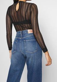 Haut noir à manches longues avec des rayures verticales transparentes, associé à un jean en denim bleu taille haute. Présente un dos à keyhole et une étiquette sur le jean.