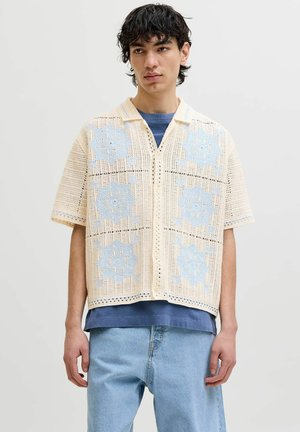 Jeune homme aux cheveux bouclés foncés portant une chemise courte en crochet beige avec des motifs floraux bleus sur un tee-shirt bleu et un jean en denim clair.