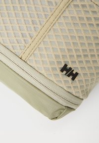 Zaino beige con motivo, superficie testurizzata, sezione inferiore verde e logo nero. Presenta una cerniera e materiale intrecciato resistente.