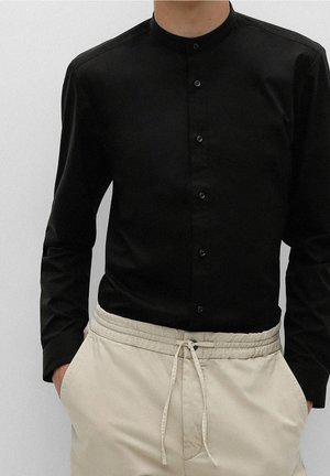 Person trägt ein schwarzes, langärmeliges Hemd mit Knopfleiste und Mandarinkragen sowie beige Hose mit Kordelzug, Hände in den Taschen.