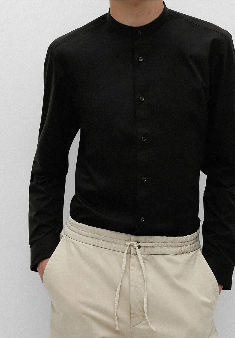 Personne portant une chemise noire à manches longues boutonnée avec un col mandarin et un pantalon beige à cordon, mains dans les poches.
