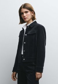 Giacca in velluto nero con due tasche sul petto, bottoni a pressione argentati e collo classico, indossata sopra una camicia bianca. Tessuto liscio, vestibilità strutturata.