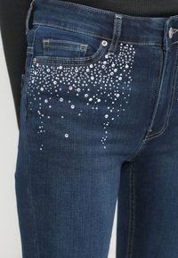 Τζιν παντελόνια από σκουρόχρωμο denim με διαβάθμιση από καθαρά στρας στην μπροστινή τσέπη και το μηρό, διακοσμημένα με ασημί εξαρτήματα.