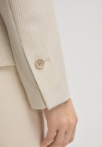 Beżowy rękaw kurtki w wzór, z teksturowanym deseniem w jodełkę; posiada okrągły beżowy guzik blisko mankietu.