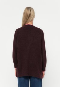 Pull en tricot bordeaux avec une finition texturée, doté de manches longues et d'un accent rose au niveau du poignet, vu de dos.