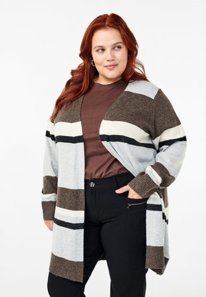 Pruhovaný cardigan v šedej, bielej a hnedej farbe, pletený materiál, otvorený predný diel, dlhé rukávy, rebrované manžety, nosený cez hnedý top a čierne nohavice.
