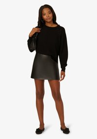 Maglione nero a maglia con dettagli intrecciati, abbinato a una mini gonna nera in ecopelle. La modella tiene una piccola borsa nera. Scarpe basse nere completano il look.