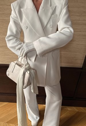 Balts, dubultpogu žakete ar teksturētu audumu, atbilstošas bikses un balti dekoratīvi cimdi. Komplekts papildināts ar nelielu gaišas krāsas rokassomu ar šalli.