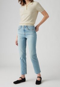 Mujer vestida con jeans rectos de color azul claro, camiseta polo de manga corta beige y zapatos de Mary Jane negros, de pie contra un fondo liso.