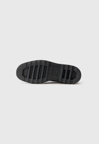 Suela de zapato de goma negra con una superficie texturizada y un patrón de relieve pronunciado, que presenta surcos profundos para tracción y estabilidad.