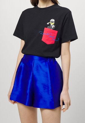 Personne portant un t-shirt noir avec une poche rouge et un motif canard cartoon, associé à un short bleu brillant taille haute.