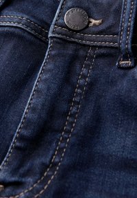 Jeans en denim bleu foncé avec un bouton en métal visible et des coutures beige contrastantes, montrant un gros plan de la texture du tissu et des coutures.