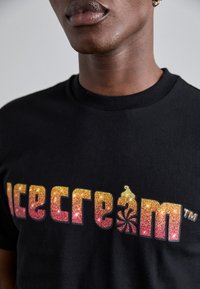 Sort bomulds T-shirt med et glitrende gradient logo, der står "icecrem" i gult til pink, med et isvaffel ikon og svømmedetalje.