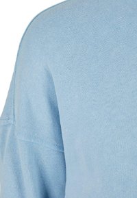 Šviesiai mėlynas megztinis su minkšta tekstūra ir laisvu siluetu, matomomis siūlėmis ir lygiu paviršiumi.