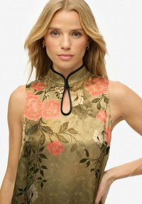 Blouse sans manches en tissu vert olive avec un motif floral comportant des fleurs roses et blanches. Col haut avec une ouverture en forme de œillet et finitions noires.