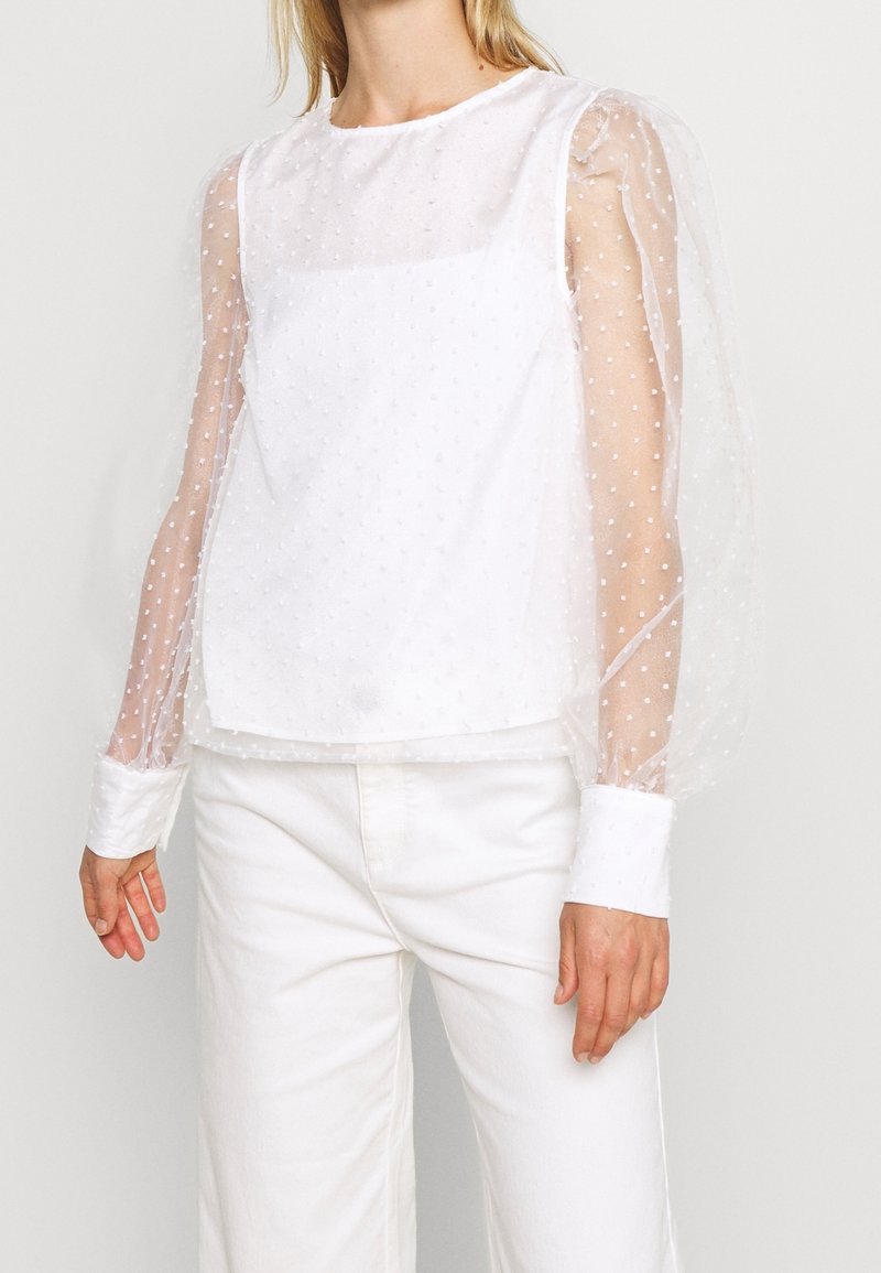 Femme portant un chemisier blanc avec des manches longues transparentes à pois et un pantalon blanc à jambes larges sur un fond clair uni.