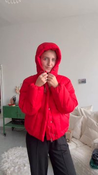 Czerwona kurtka puchowa z kapturem, z pikowaną fakturą, frontowymi guzikami i lekko oversize'owym krojem. Noszona przez postać trzymającą kołnierz.