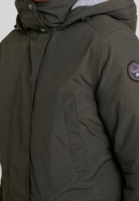 Donkergroene geïsoleerde winterjas met een capuchon, twee voorzakken, knoopsluiting en een rond logopatch op de bovenste mouw.