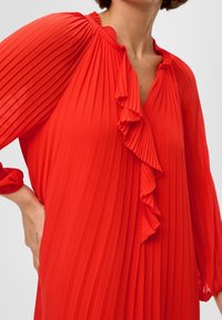 Rotes, gefaltetes Kleid mit einem fransigen Ausschnitt und weiten Ärmeln. Der strukturierte Stoff verleiht Tiefe; das Design besteht aus sanften Drapierungen.
