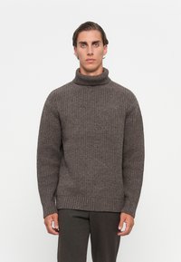 Ung man med slickat hår iförd en brun ribbad polotröja och mörka byxor, stående framför en vit bakgrund.