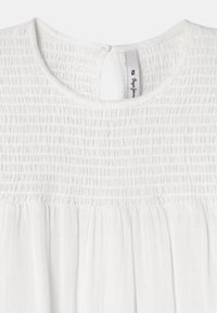 Bílá blůza s nařasenou texturou na výstřihu. Má kulatý výstřih, užší zadní zapínání a lehký, splývavý materiál.