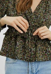 Blusa verde con motivo floreale, con volant e vita arricciata, dotata di maniche lunghe e scollatura a V. Jeans di denim chiaro sono parzialmente visibili.