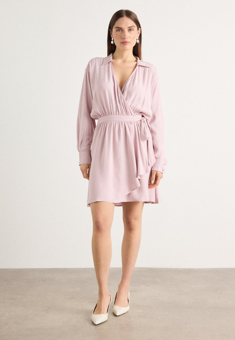 Wrap dress in light pink fabric with a deep V-neck, long sleeves, and a tie waist. Features a ruffled hem and vertical stripe texture.