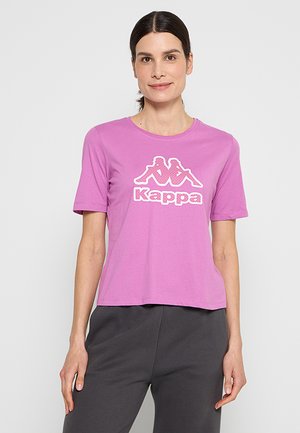 Donna con capelli scuri che indossa una maglietta rosa Kappa e pantaloni grigio scuro, in piedi davanti a uno sfondo grigio chiaro uniforme.