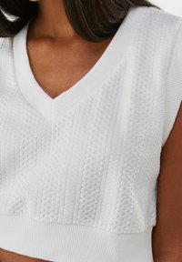 Weißer, kurz geschnittener Pullover mit strukturiertem Zopfmuster, V-Ausschnitt und gerippten Akzenten entlang der Kanten.