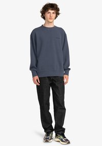 Felpa grigia con scollo a girocollo e polsini a coste, abbinata a jeans neri e sneakers multicolori. Accento di logo semplice sul petto.