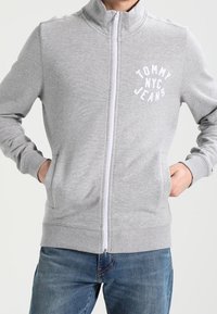 Lichtgrijze zip-up sweatshirt met hoge kraag, voorzien van een witte "TOMMY NYC JEANS" logo op de borst en ribgebreide manchetten en zoom.