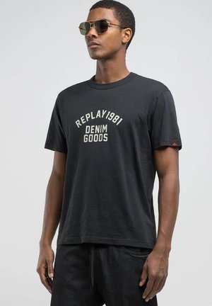 Camiseta negra de manga corta con "REPLAY1981 DENIM GOODS" impreso en blanco, cuello redondo y un ajuste relajado. Combinada con pantalones oscuros y gafas de sol.
