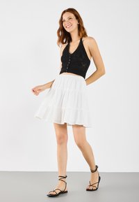 Top tricotat scurt, negru, cu nasturi, asortat cu o fustă albă fluidă, cu volane. Modelul poartă sandale negre cu barete. Texturi netede și contrast ușor.
